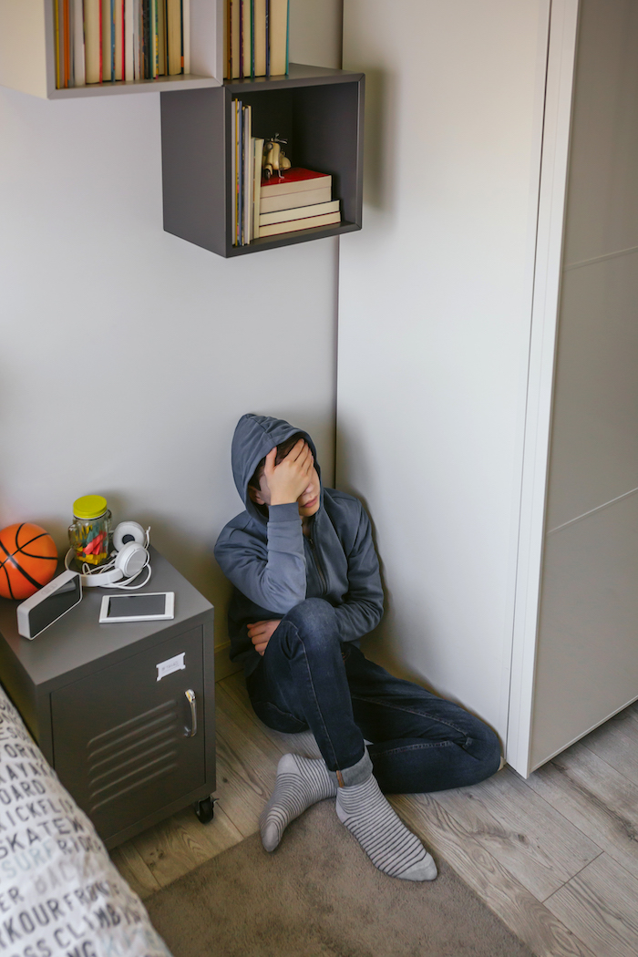 Unrecognizable depressed male teenager with his hand on his head sitting on the floor in his bedroom