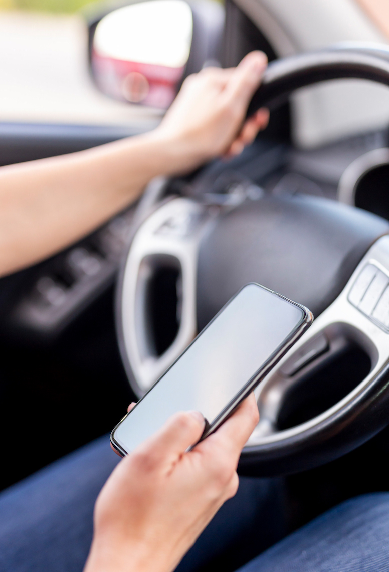 Detail of female hands holding a steering wheel and typing a text message on a smart phone while driving a car