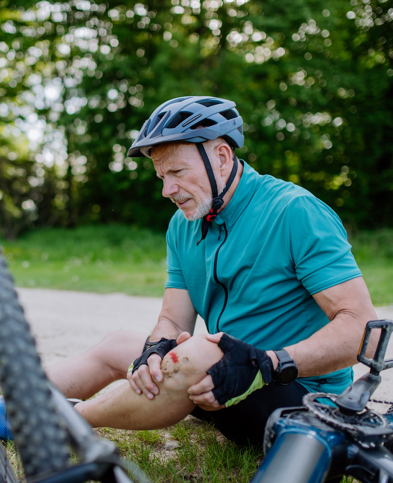 An active senior man in sportswear fell off bicycle on the ground and hurt his knee, in park in summer.
