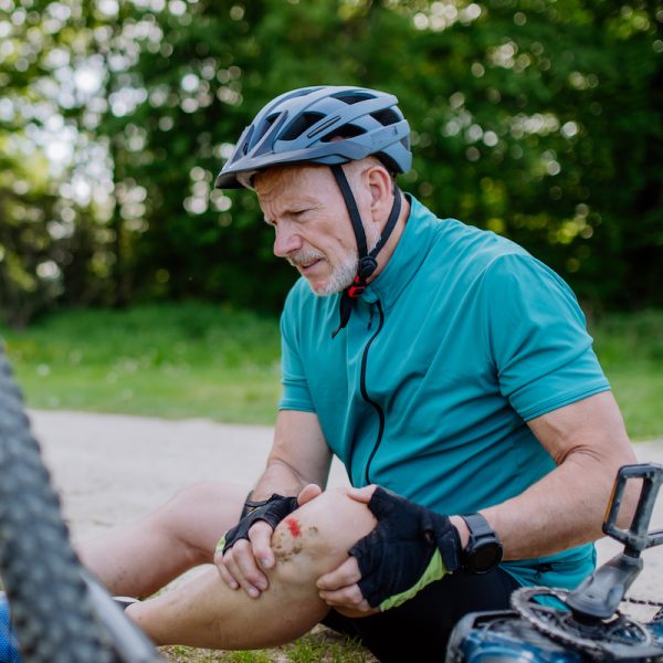 An active senior man in sportswear fell off bicycle on the ground and hurt his knee, in park in summer.