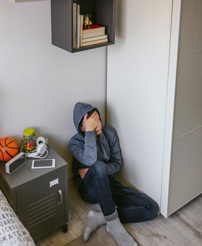 Unrecognizable depressed male teenager with his hand on his head sitting on the floor in his bedroom