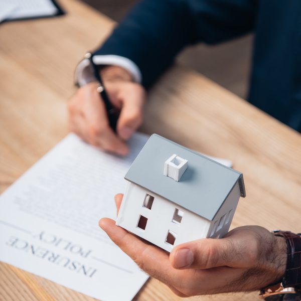 cropped view of lawyer holding house model near documents with insurance policy lettering on table