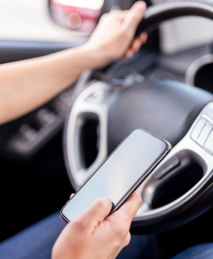 Detail of female hands holding a steering wheel and typing a text message on a smart phone while driving a car